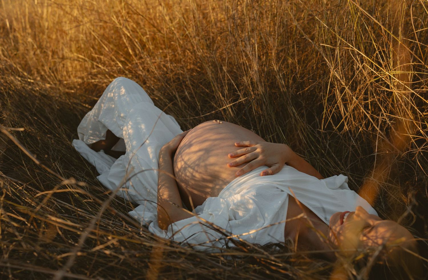 A smiling baby in a white onesie lying on a white dress while being tickled in warm, sepia-toned lighting.