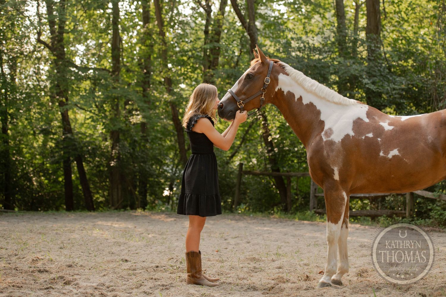 A person in a black dress pets a brown and white paint horse in a wooded outdoor setting.