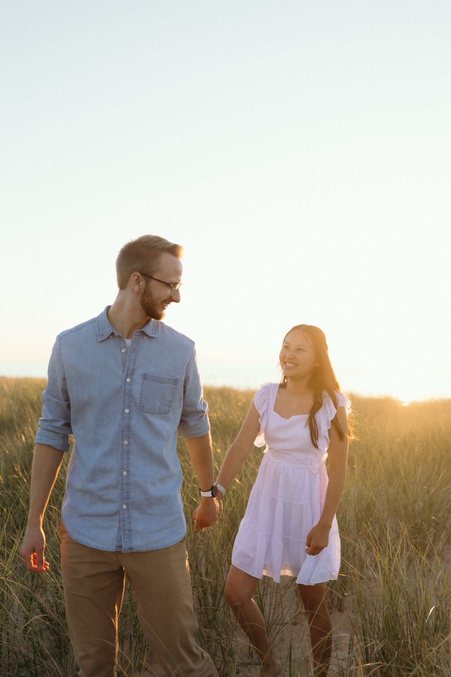 A couple laughs together in a grassy field during golden hour sunset, wearing casual summer outfits.