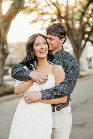 A romantic couple embraces and laughs together outdoors during golden hour on a tree-lined path.