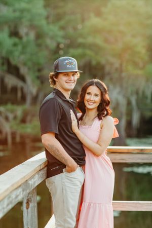 A couple poses on a wooden dock at sunset, with her wearing a pink dress and him in a baseball cap and dark polo.