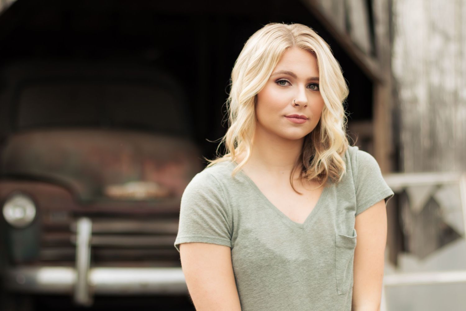 Woman in sage green v-neck t-shirt poses outdoors with soft blonde wavy hair in casual portrait setting.