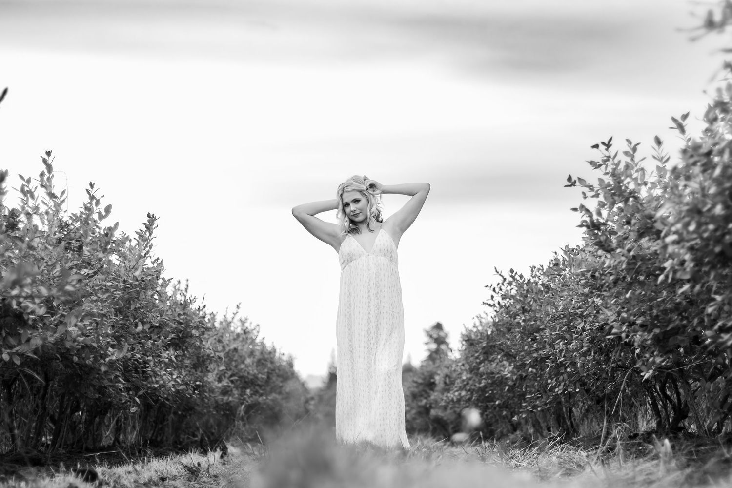 A figure in a flowing white dress stands between two rows of blooming orchard trees in a dreamy black and white scene.
