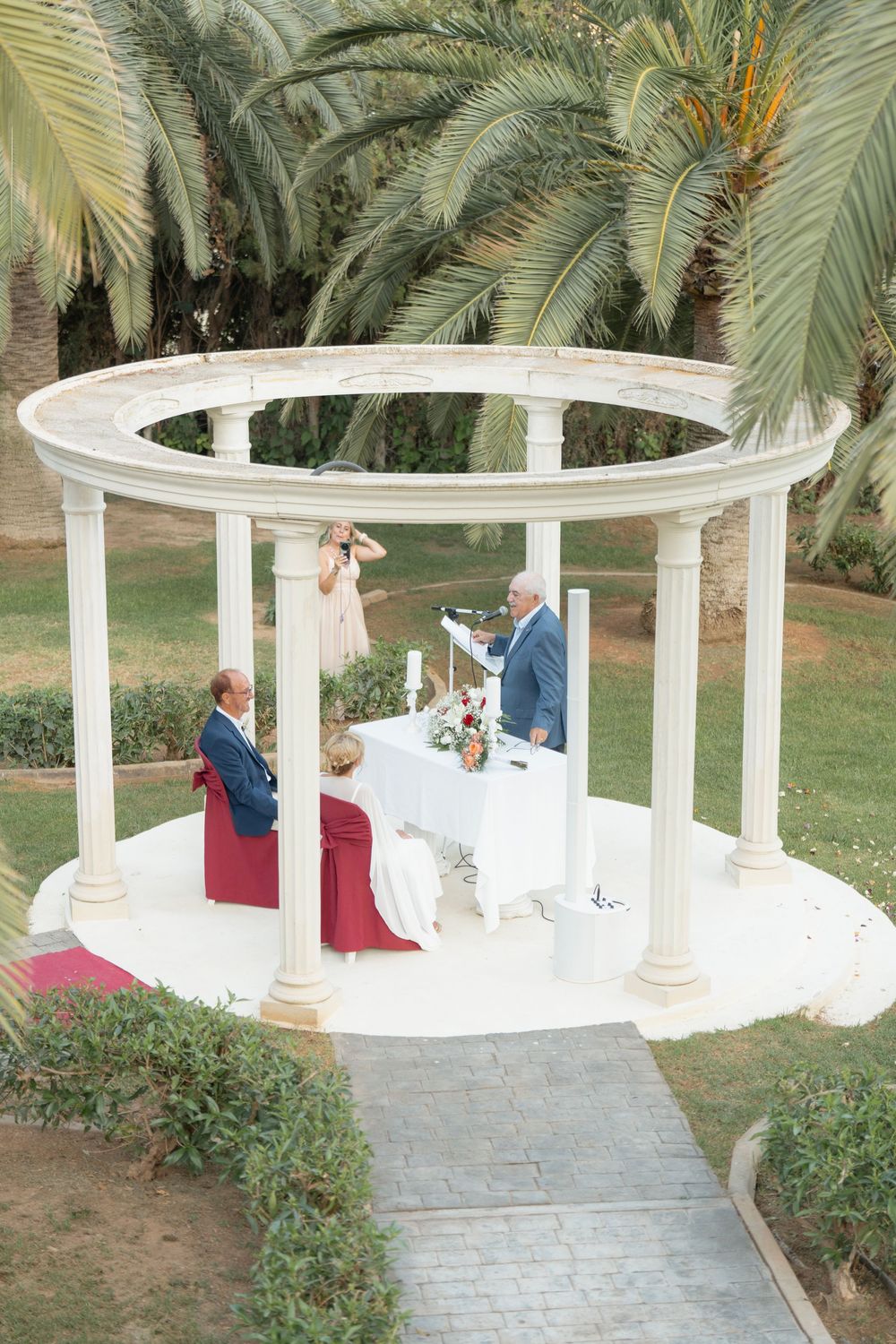 Elegant outdoor wedding ceremony in a white circular gazebo surrounded by lush palm trees and manicured gardens.