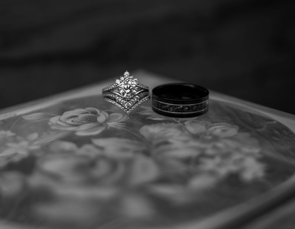 Black and white close-up of elegant diamond wedding ring set and black groom band on floral surface.