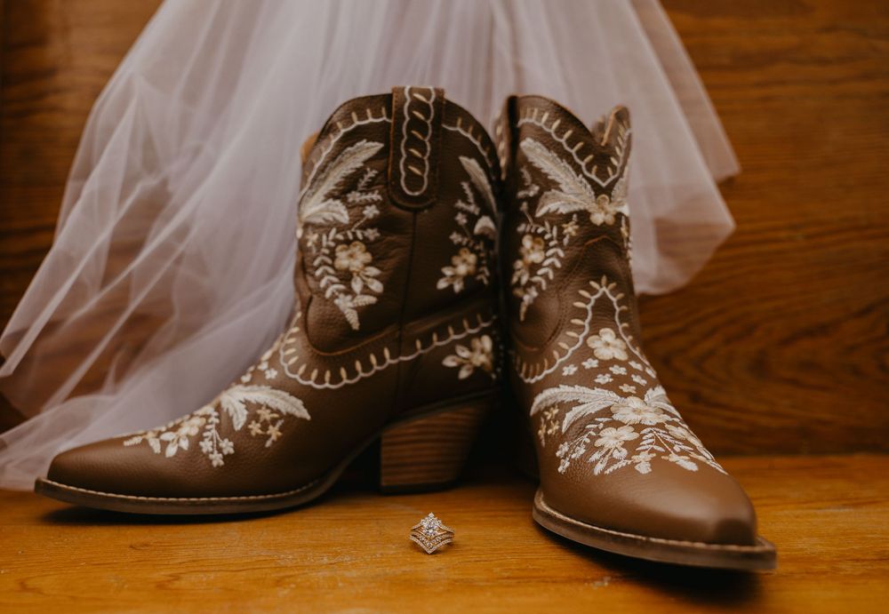 Embroidered brown cowboy boots with white floral design beside a bridal veil and diamond ring.