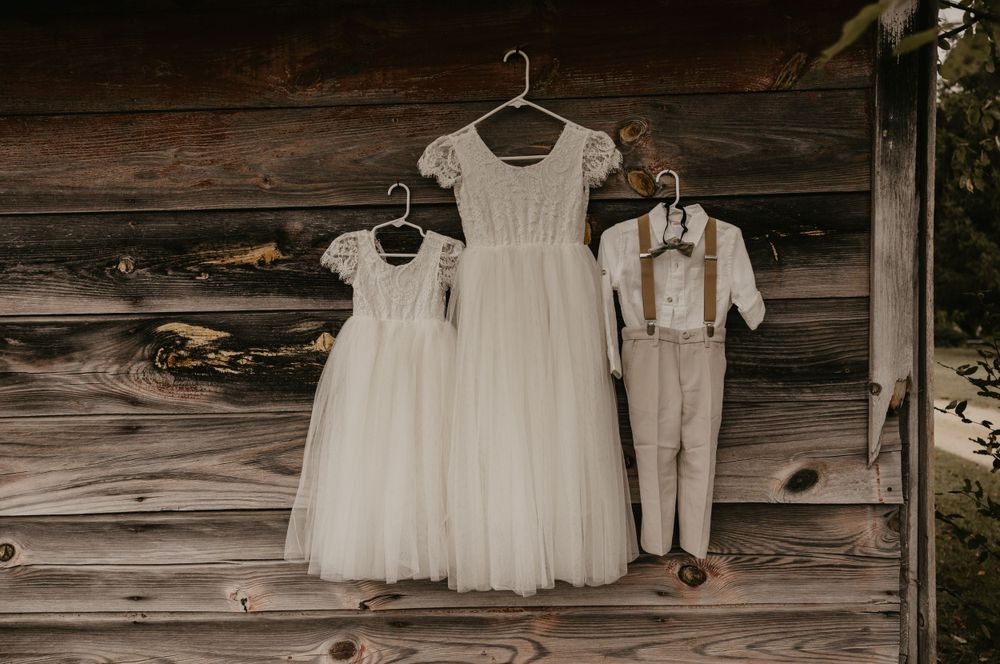 Two white flower girl dresses and ring bearer outfit hanging on rustic wooden barn wall for wedding.