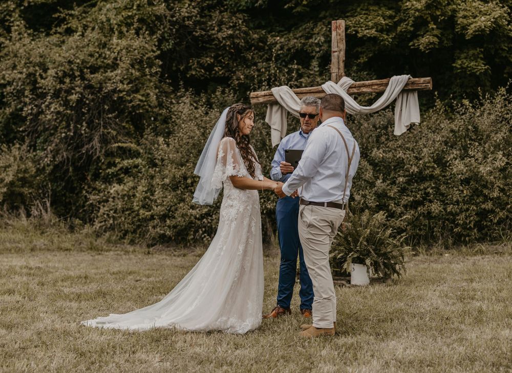 Bride and groom exchange vows at rustic outdoor wedding ceremony beneath wooden arch draped with white fabric.