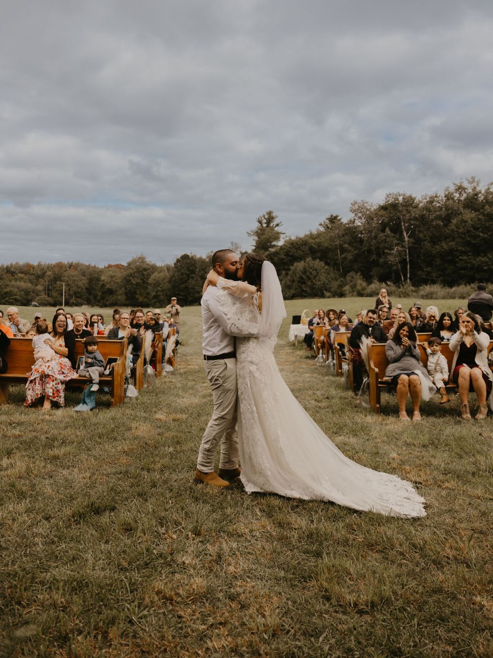 Bride and groom share first kiss at outdoor wedding ceremony with guests seated on wooden pews in field.
