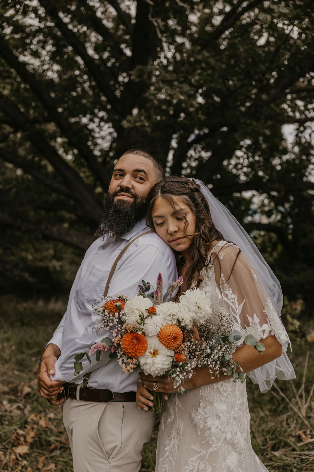 Bearded groom in suspenders embraces bride holding orange and white floral bouquet under large tree.