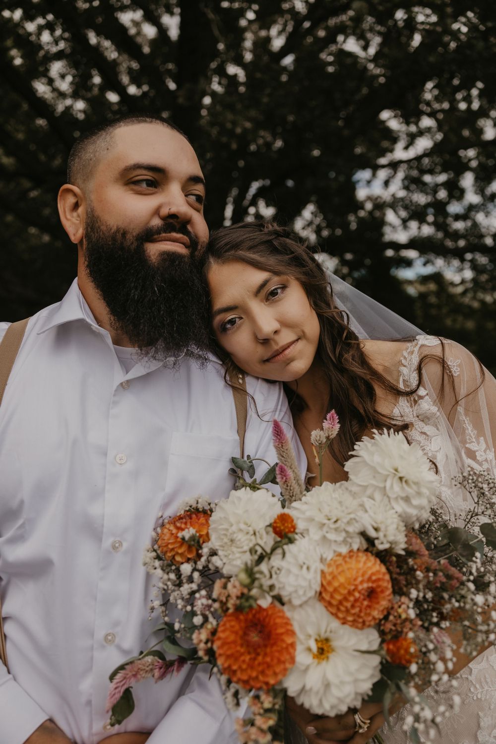 Bride rests head on bearded grooms shoulder holding vibrant orange and white dahlia wedding bouquet outdoors.