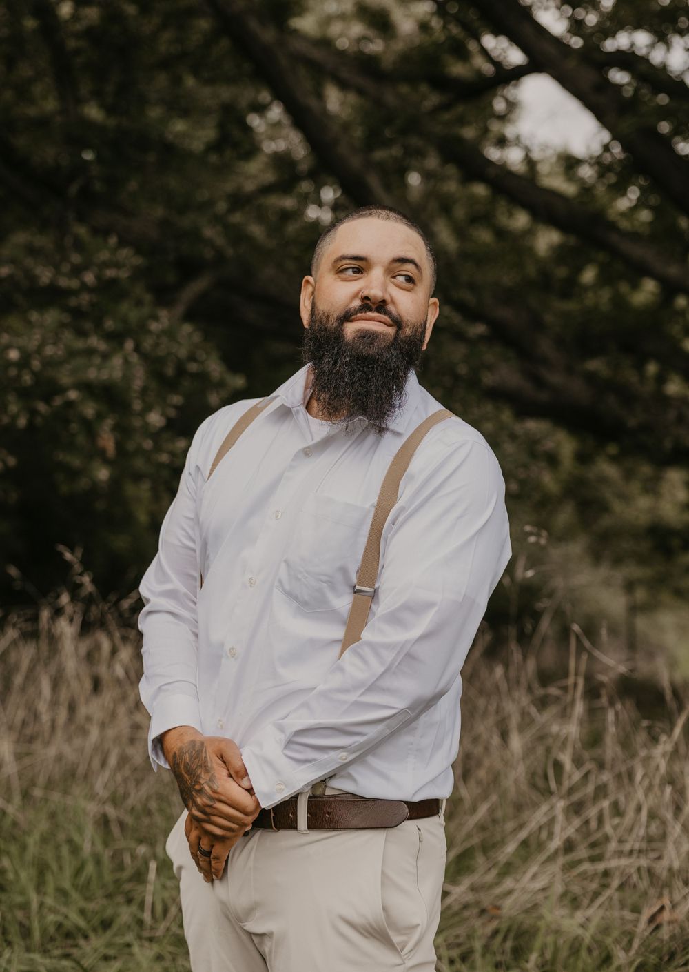 Bearded groom in white shirt and tan suspenders poses confidently in rustic outdoor field setting.