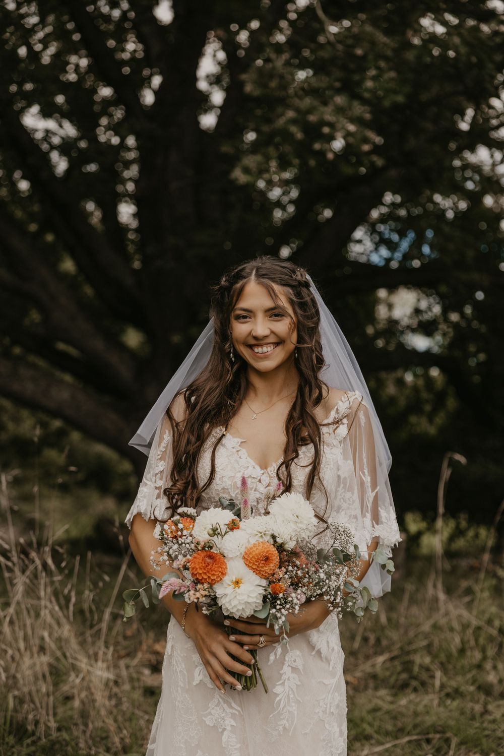 Smiling bride in lace gown and veil holds orange and white dahlia bouquet in wooded outdoor setting.