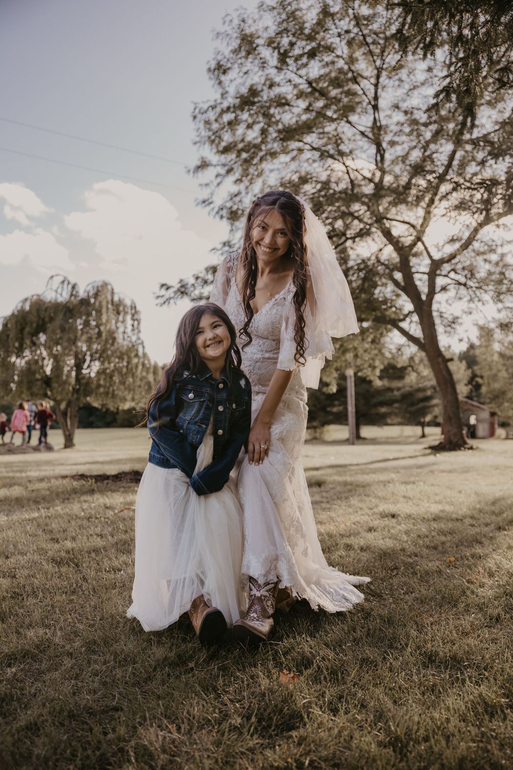 Smiling bride in lace gown kneels beside young flower girl in denim jacket and white dress outdoors.