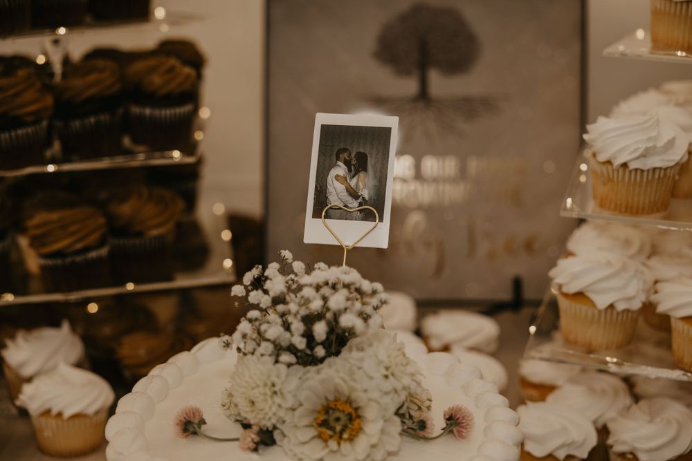 Wedding dessert table features Polaroid photo topper on white floral arrangement surrounded by cupcakes.