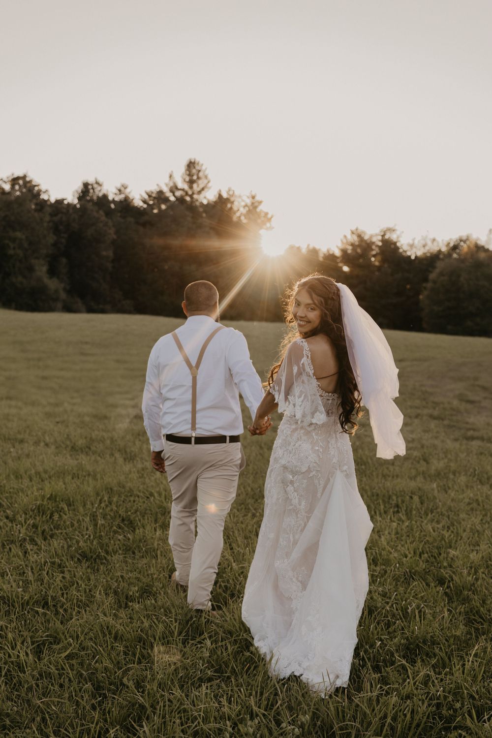 Bride and groom walking hand in hand through a sunlit green field at golden hour sunset.