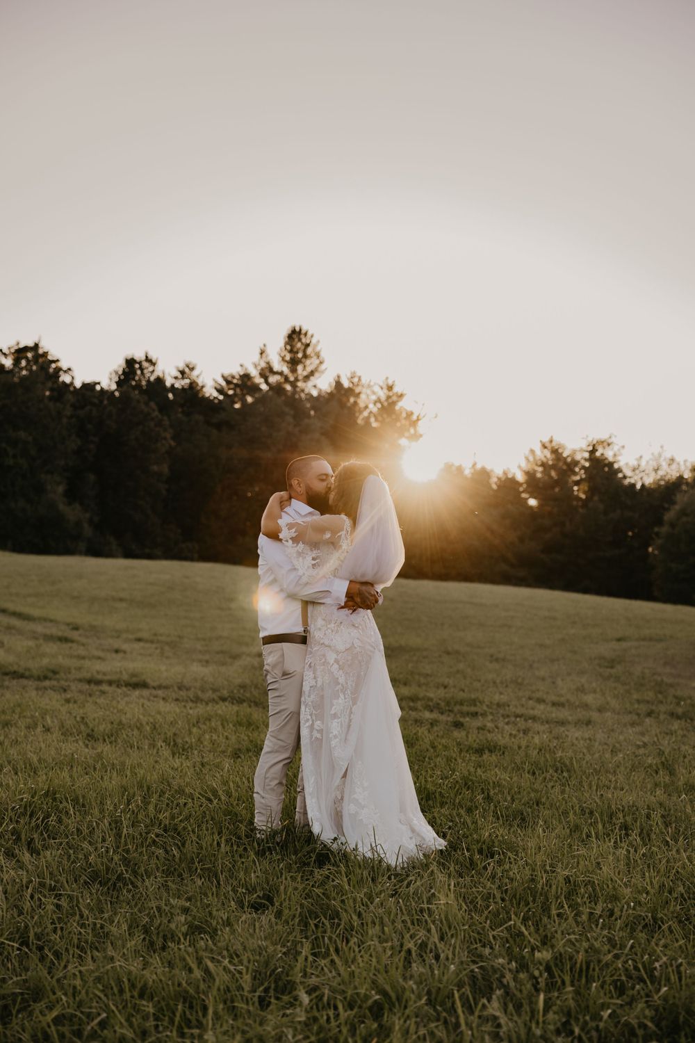 Bride and groom sharing a romantic embrace in a green meadow with glowing sunset behind them.