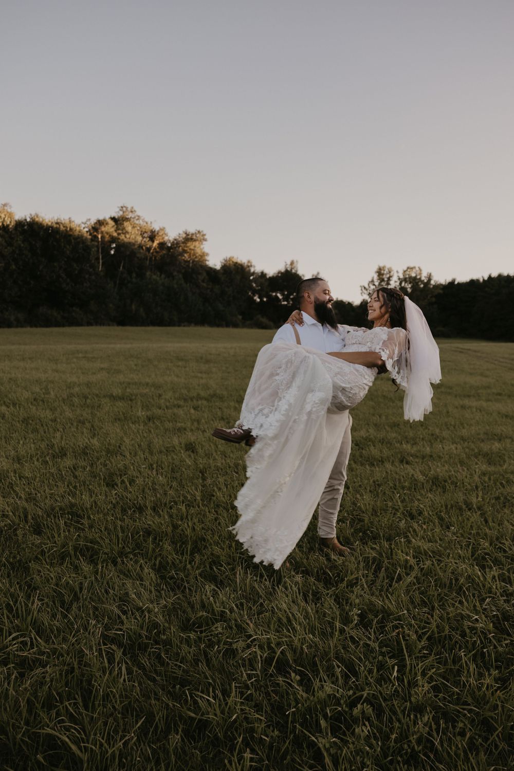 Groom lifting bride in a lace wedding gown in an open green field at dusk.
