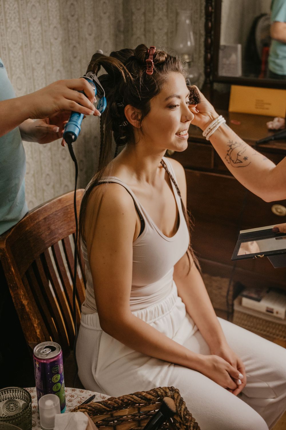 Bride getting hair and makeup done by two stylists while seated in a vintage chair.