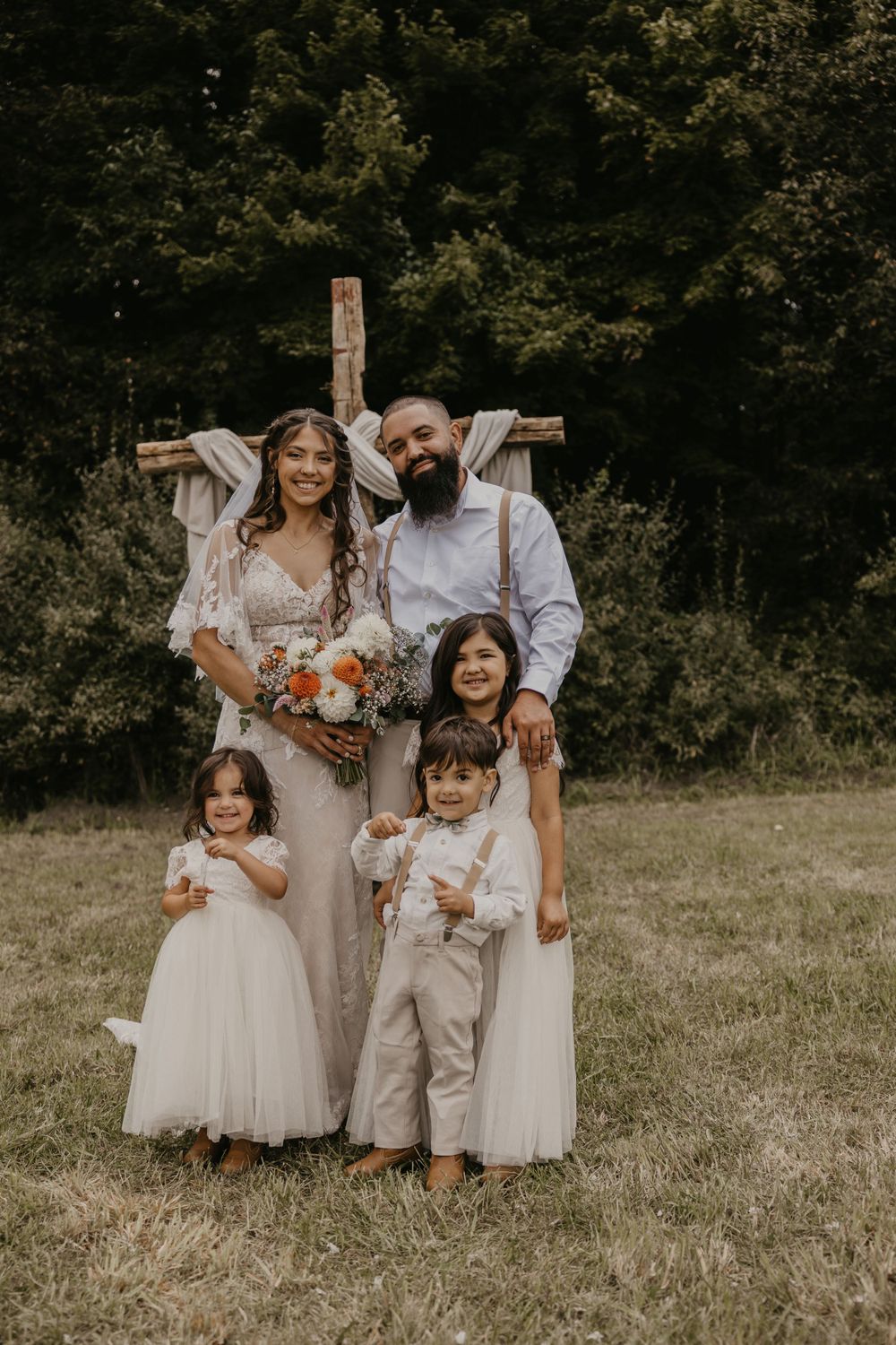 Happy wedding couple posing with three young children in front of a rustic wooden cross outdoors.