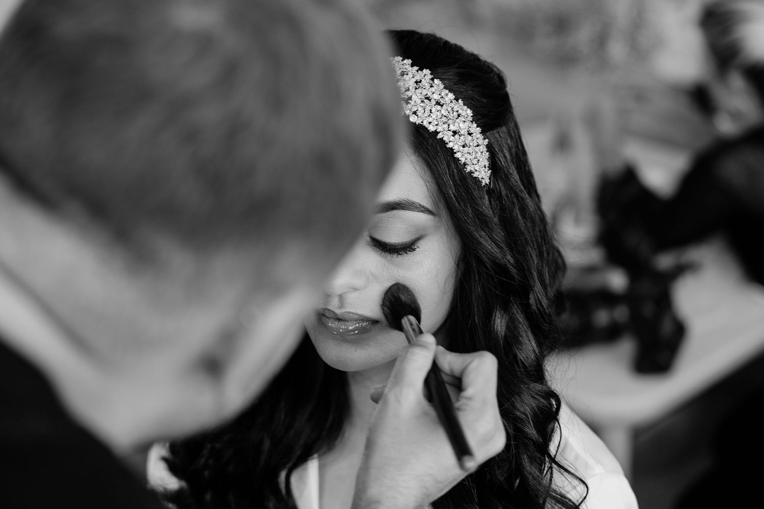 Black and white photo of makeup application during bridal preparation with sparkly headpiece detail.