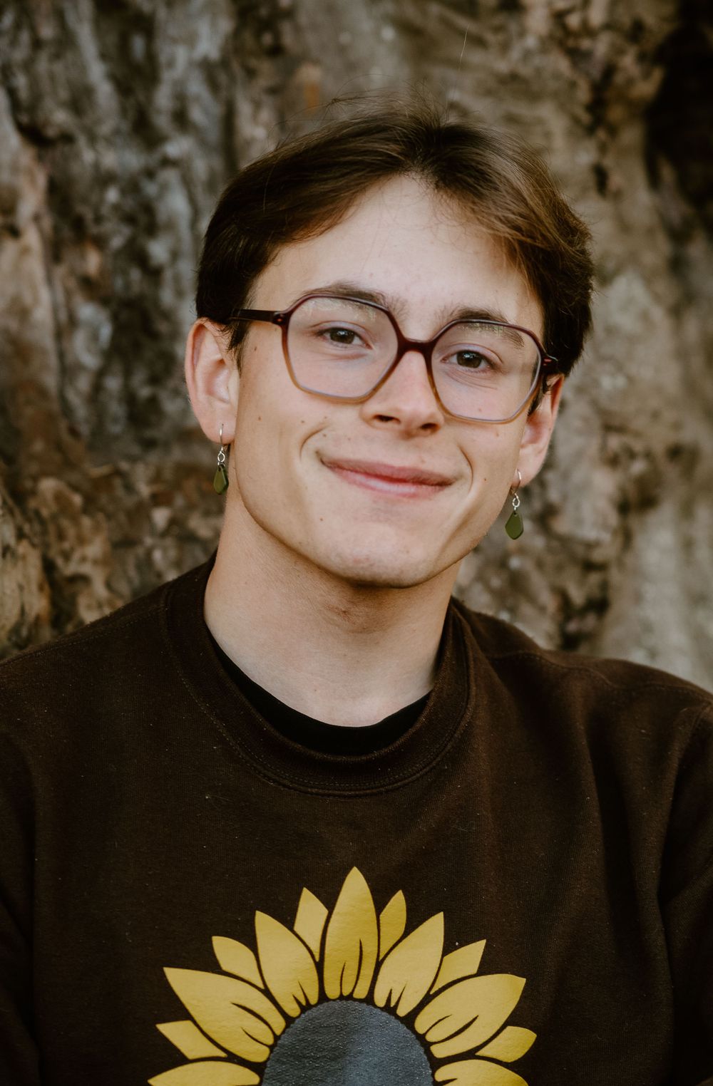 Portrait of someone wearing glasses and a brown sweater with sunflower design against tree bark.