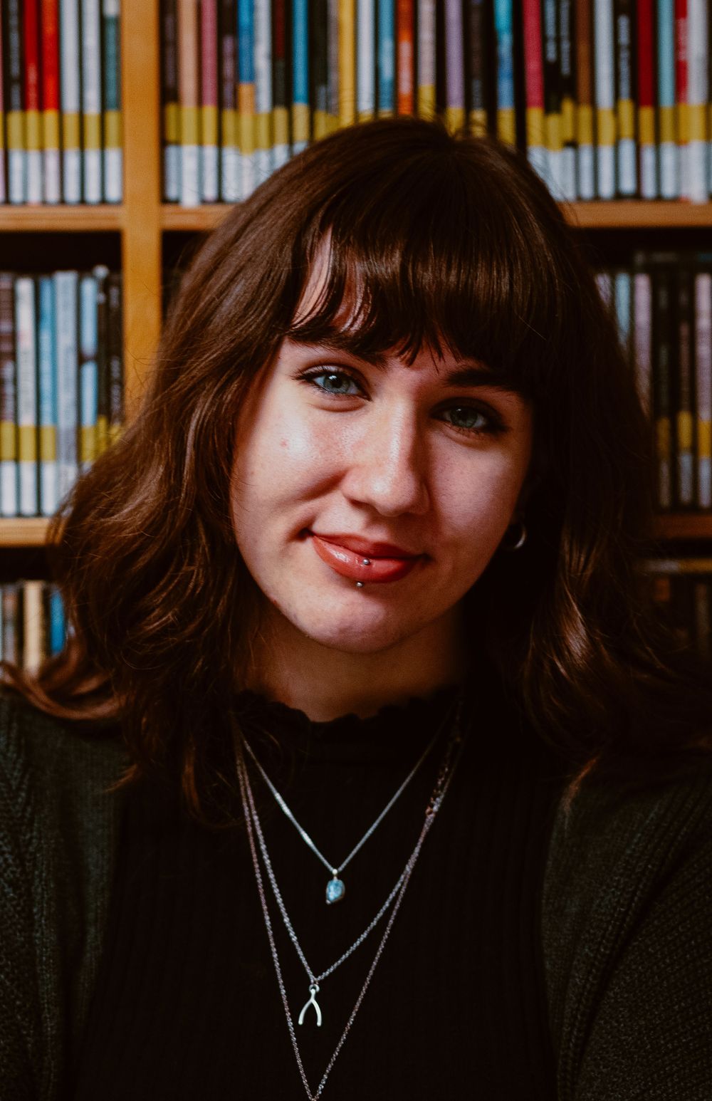 Close-up portrait in front of library shelves wearing layered necklaces and dark sweater.