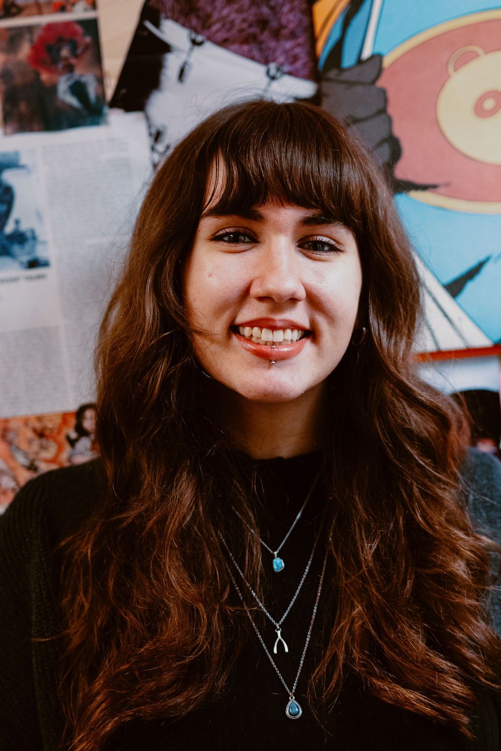 Portrait against colorful wall art wearing black top and layered pendant necklaces.