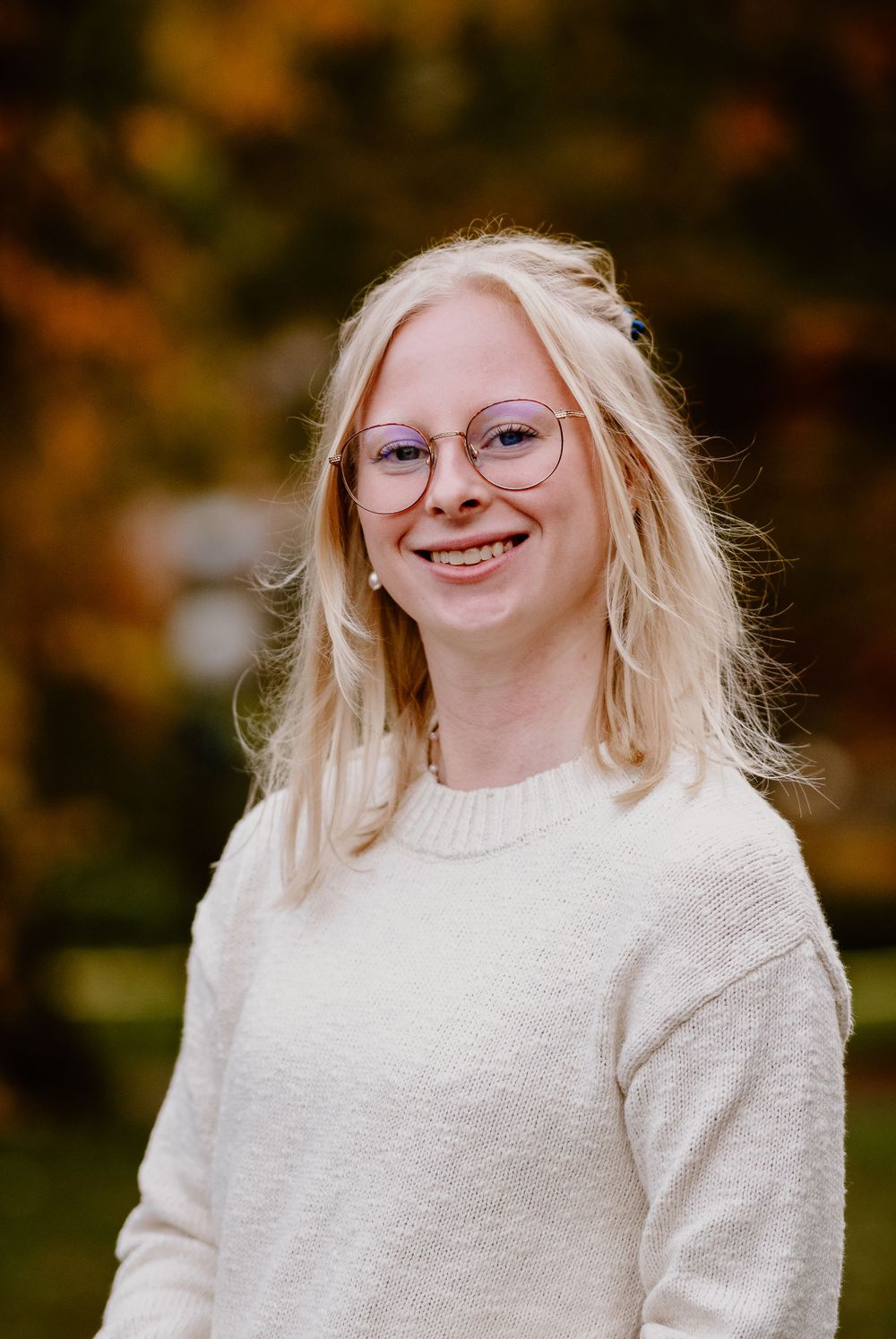 A person in round glasses and white sweater smiles warmly during an outdoor photoshoot.