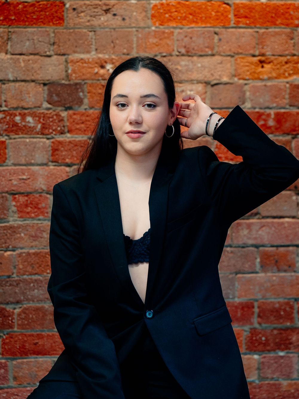 Portrait against red brick wall wearing an oversized black blazer in moody lighting.
