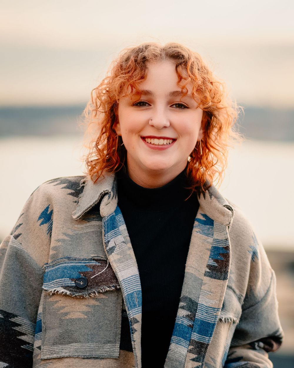 A curly redheaded person wearing a denim jacket smiling outdoors in soft natural lighting.