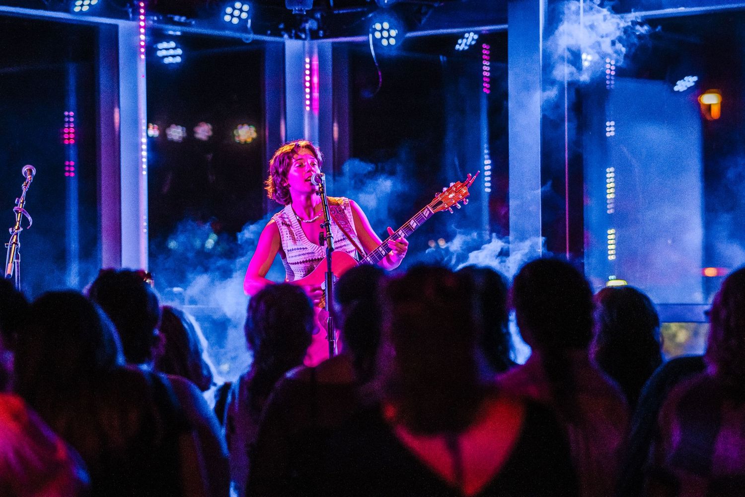 Concert performer plays guitar on stage with dramatic pink lighting and smoke effects.
