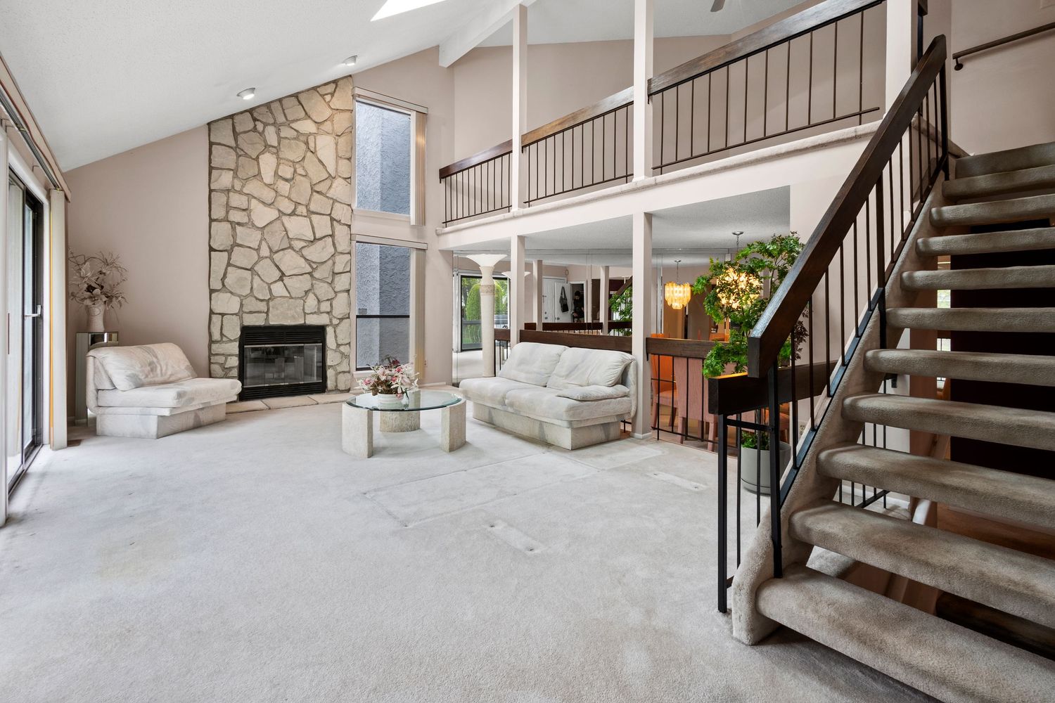 Modern living room features stone fireplace, white carpeting, floating stairs and open floor plan with tall cathedral ceiling.