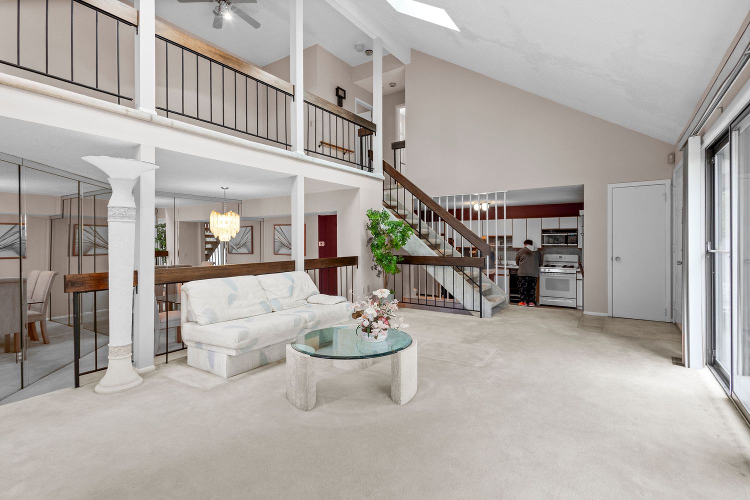 Contemporary living room with white sofa, glass table, vaulted ceiling, and loft area featuring gray carpeting and natural light.