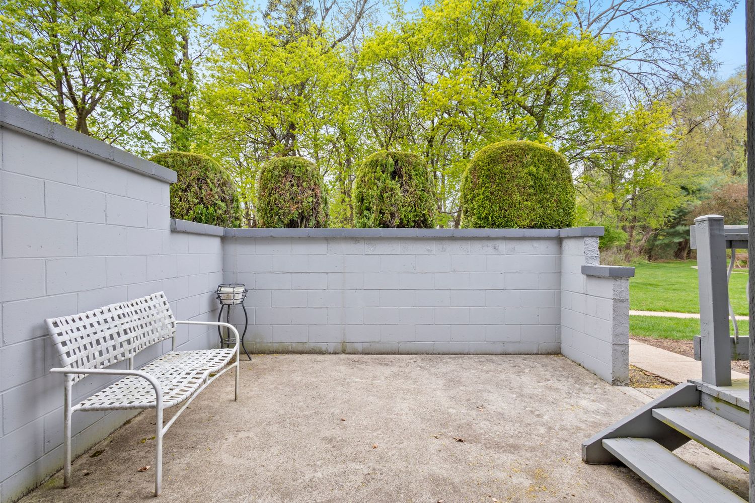 A sleek curved metal bench sits against a grey concrete retaining wall in a park-like setting with spring foliage.