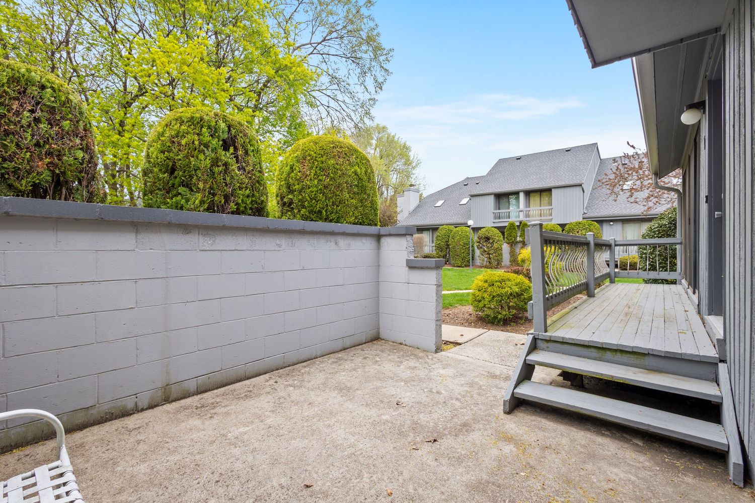A modern backyard patio area with concrete floor, white walls, wooden steps and lush green trees in the background.