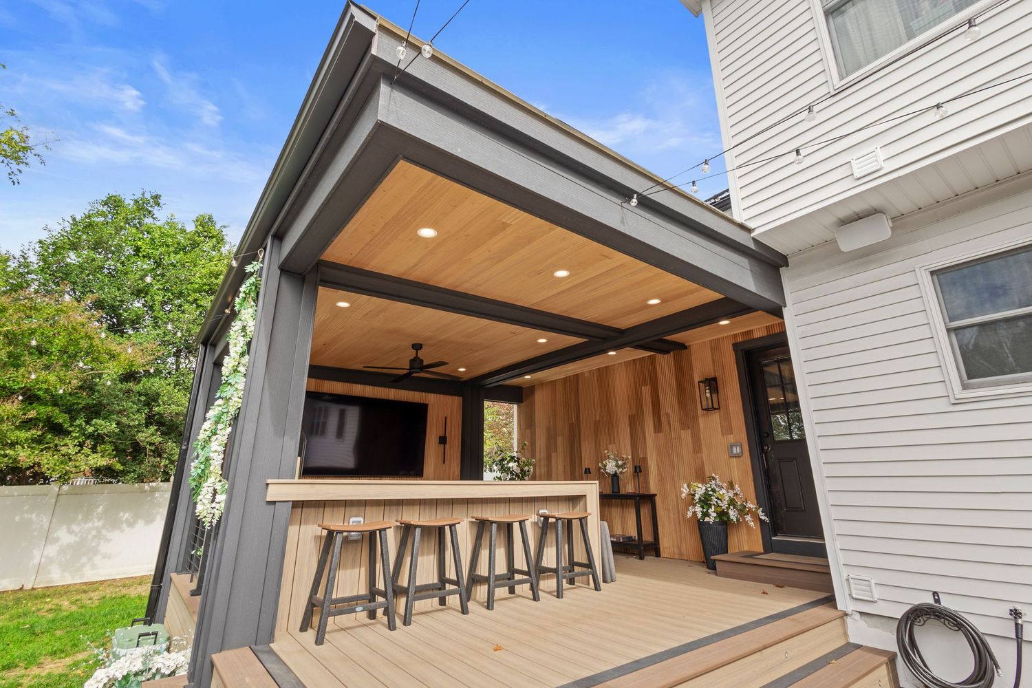 Modern outdoor deck with pergola, bar seating, wood ceiling, recessed lighting, and TV entertainment area.