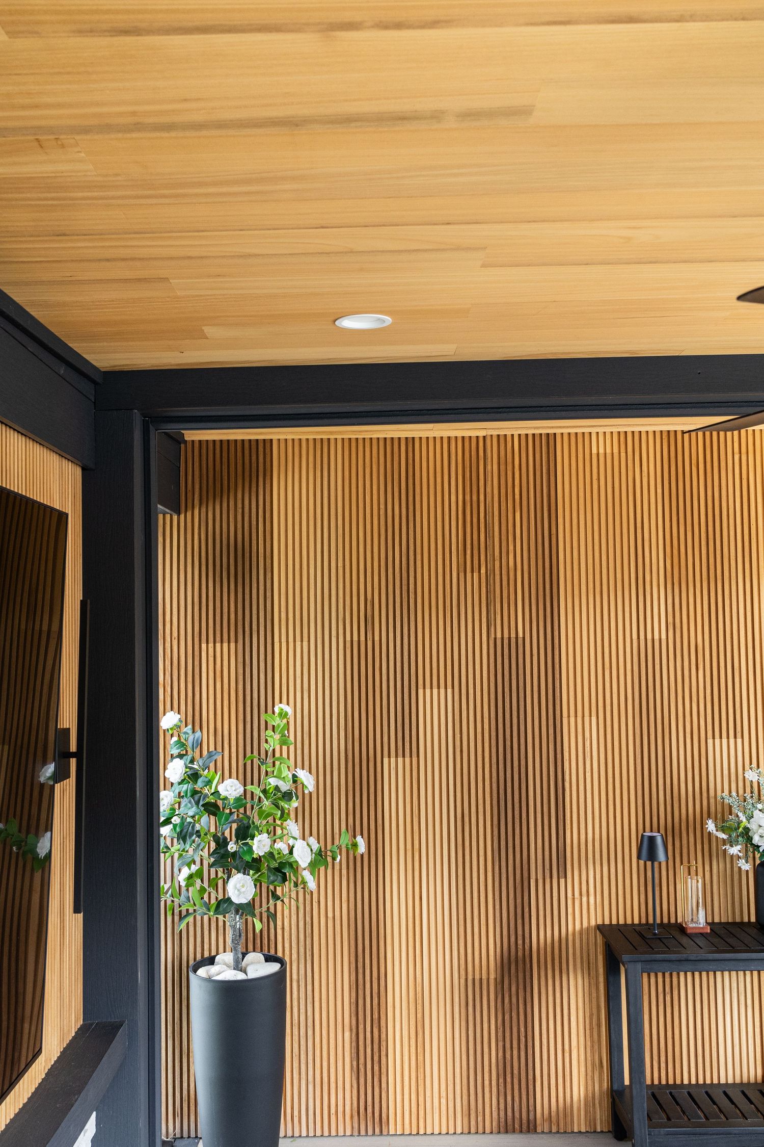 Modern interior with wooden slat wall, matching ceiling, black framing, and a tall planter with white flowers.