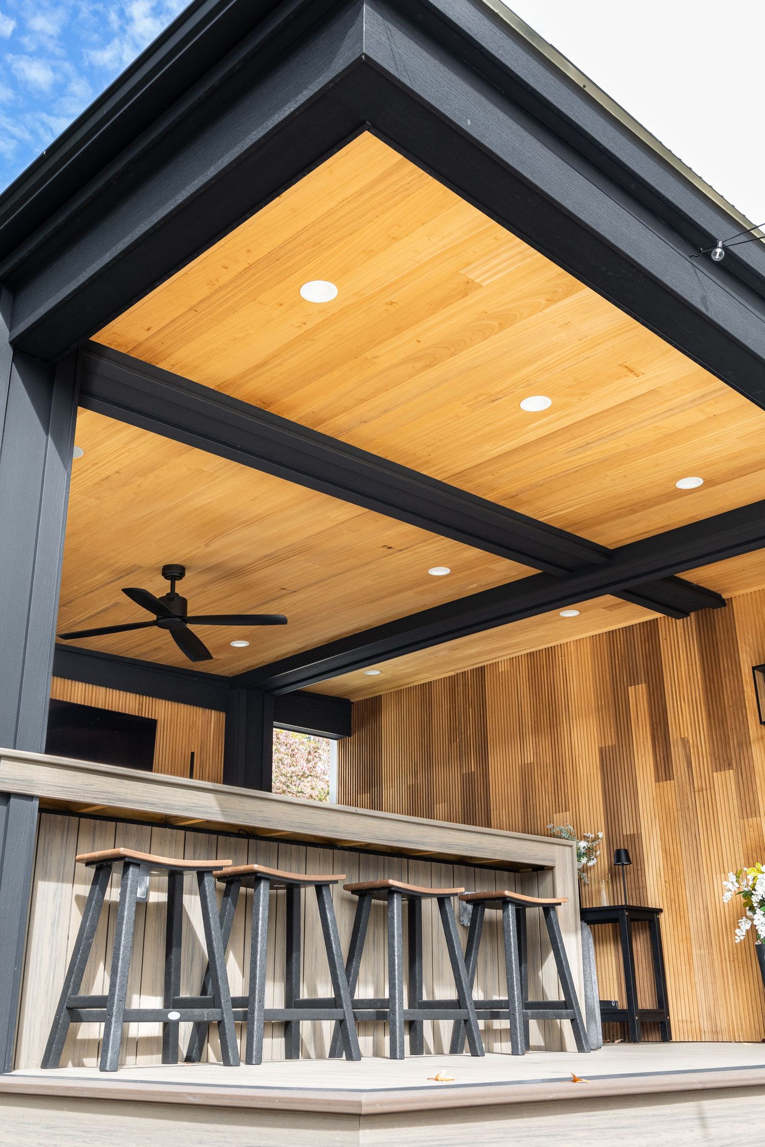 Modern outdoor covered patio bar with cedar wood ceiling, black steel beams, recessed lighting, ceiling fan, and metal bar stools.