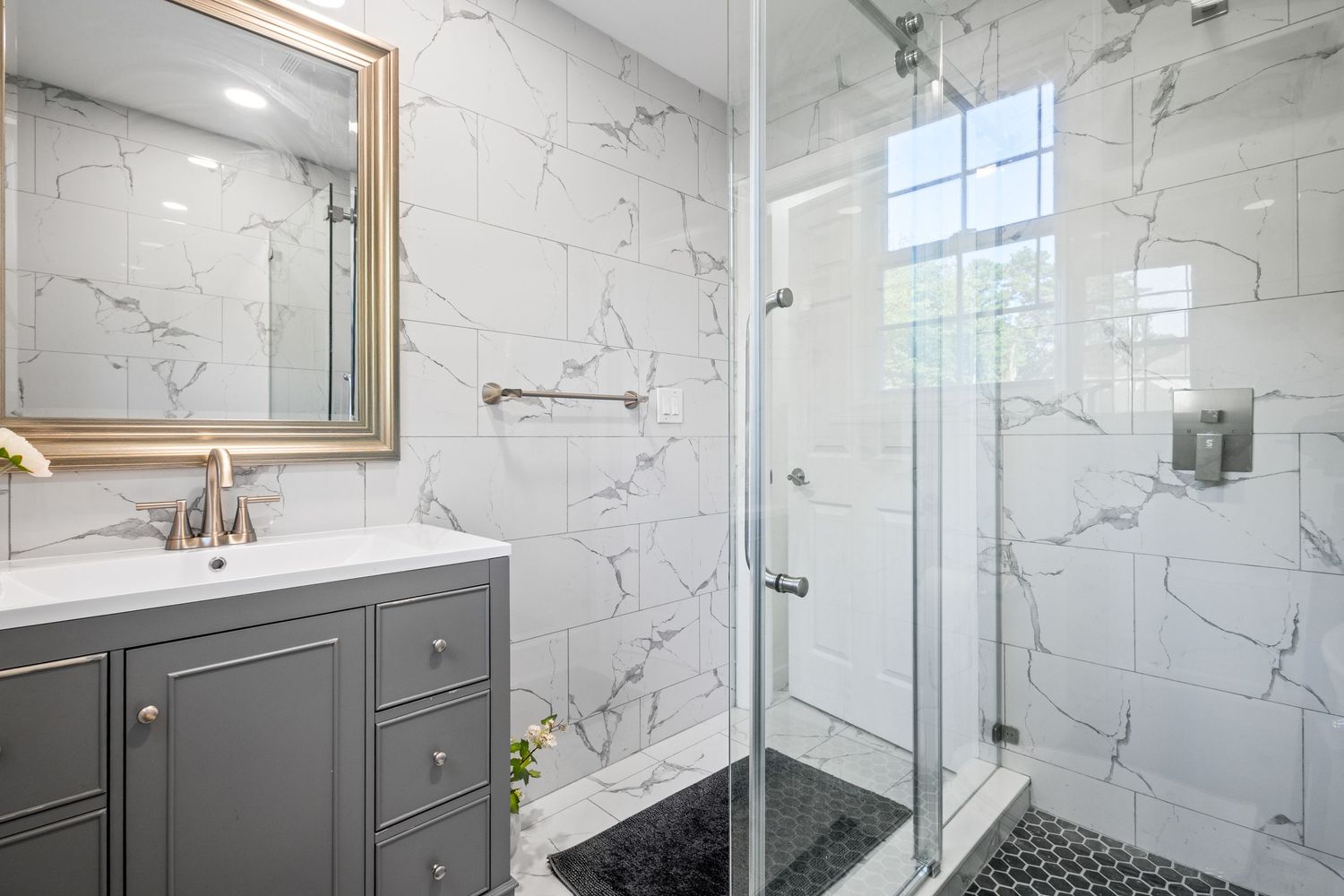 Modern bathroom interior with marble walls, gray vanity cabinet, framed mirror and glass shower enclosure.