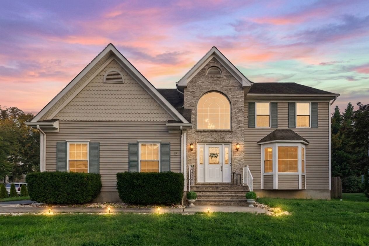 Two-story suburban home with gray siding, brick accents, arched entry, and bay window at twilight with colorful sky.