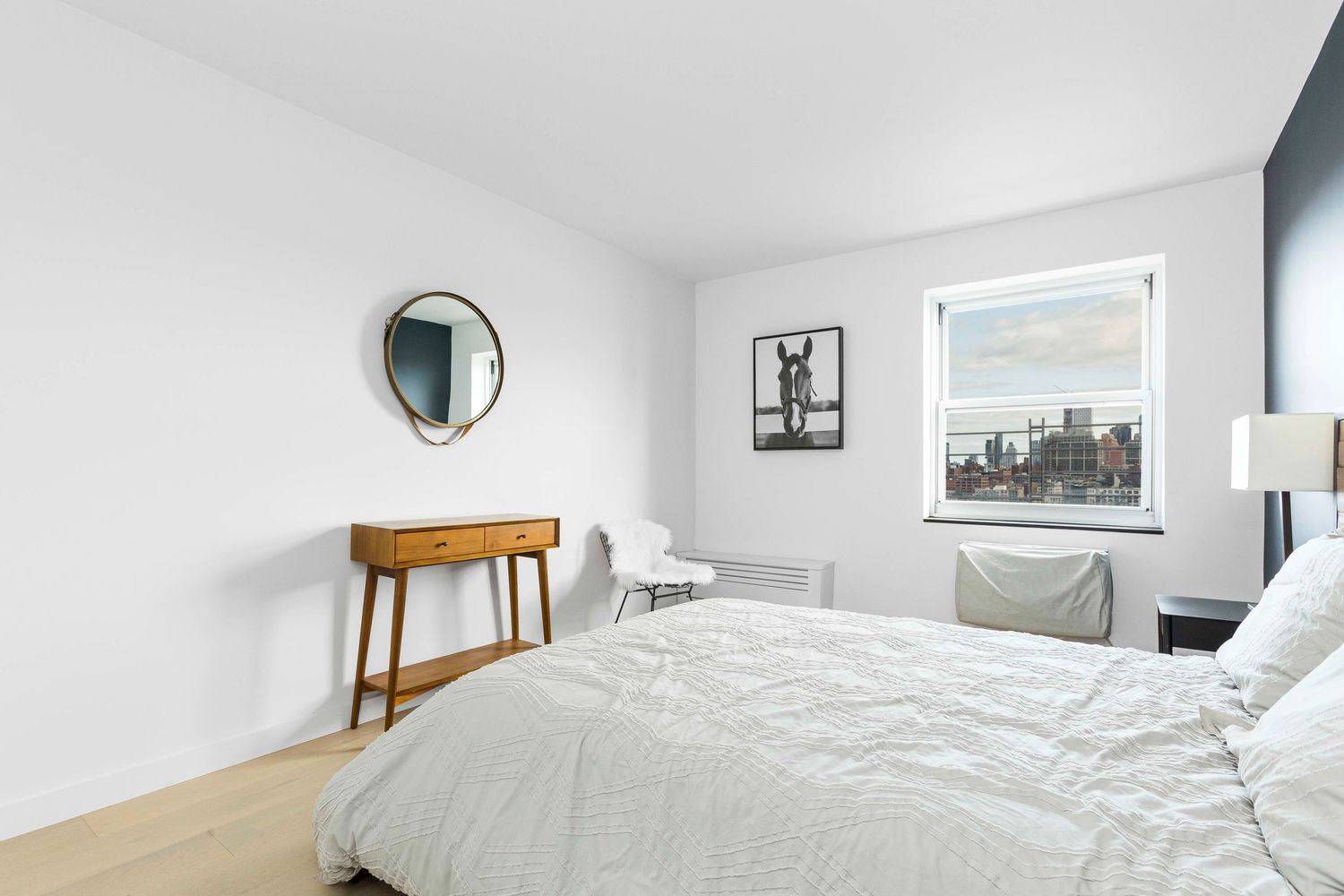 Modern bedroom with wooden console table, round mirror, and city view window.