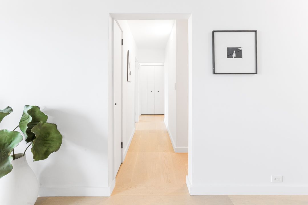 White hallway with light wood flooring, framed artwork, and potted plant.