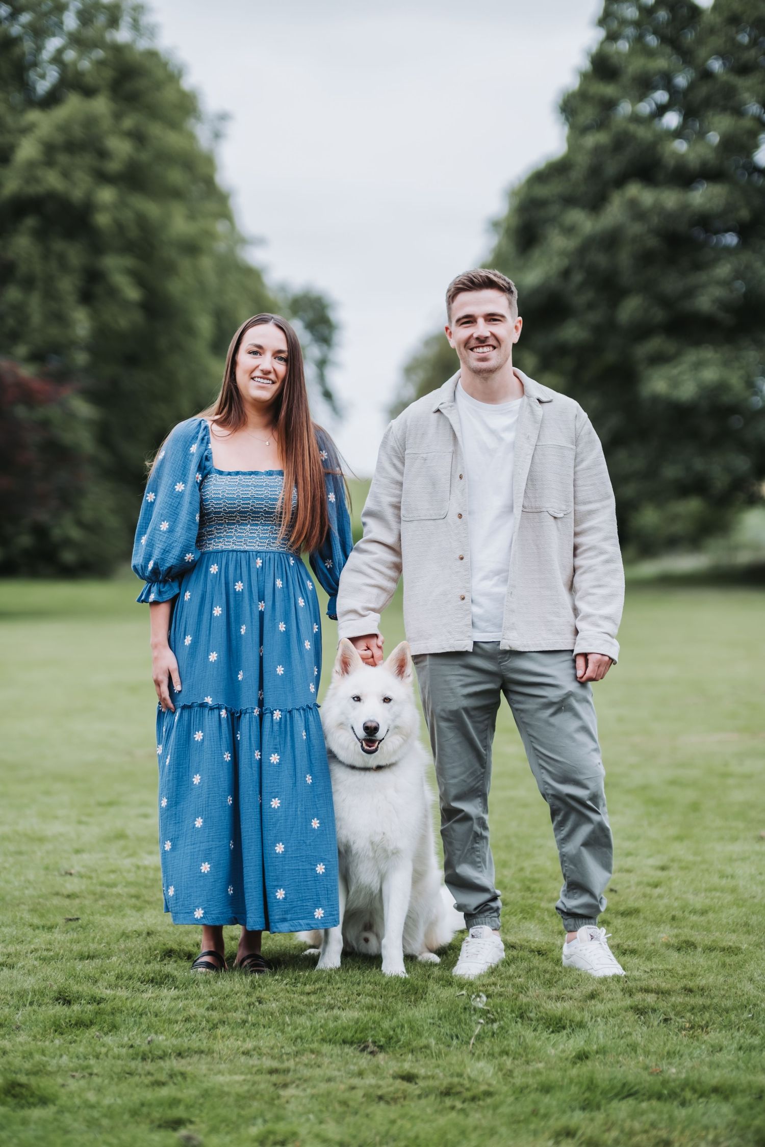 A smiling couple stands in a lush green park with their white Husky dog on a cloudy day.