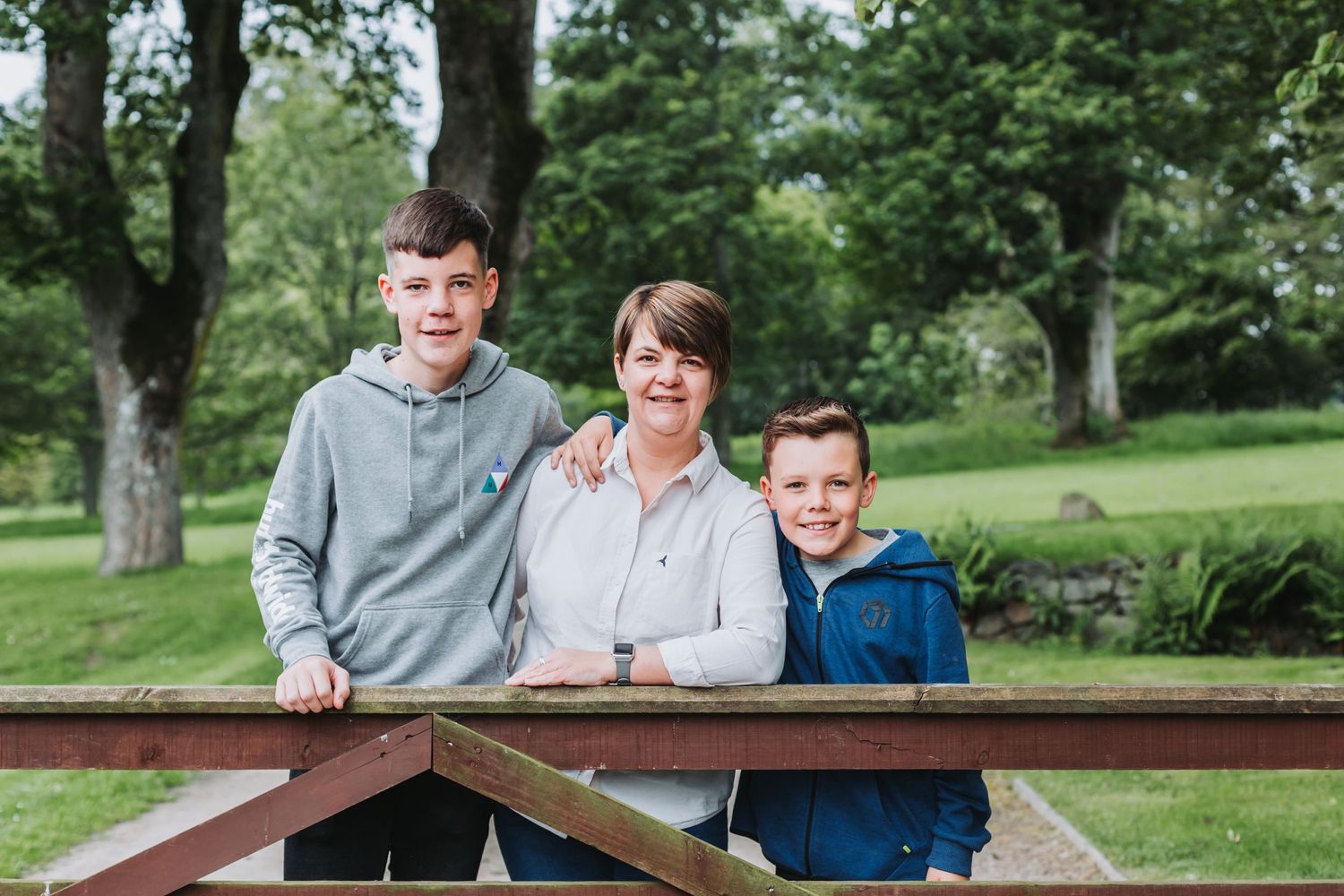A mother smiles warmly between her two sons while leaning on a wooden fence in a green park.