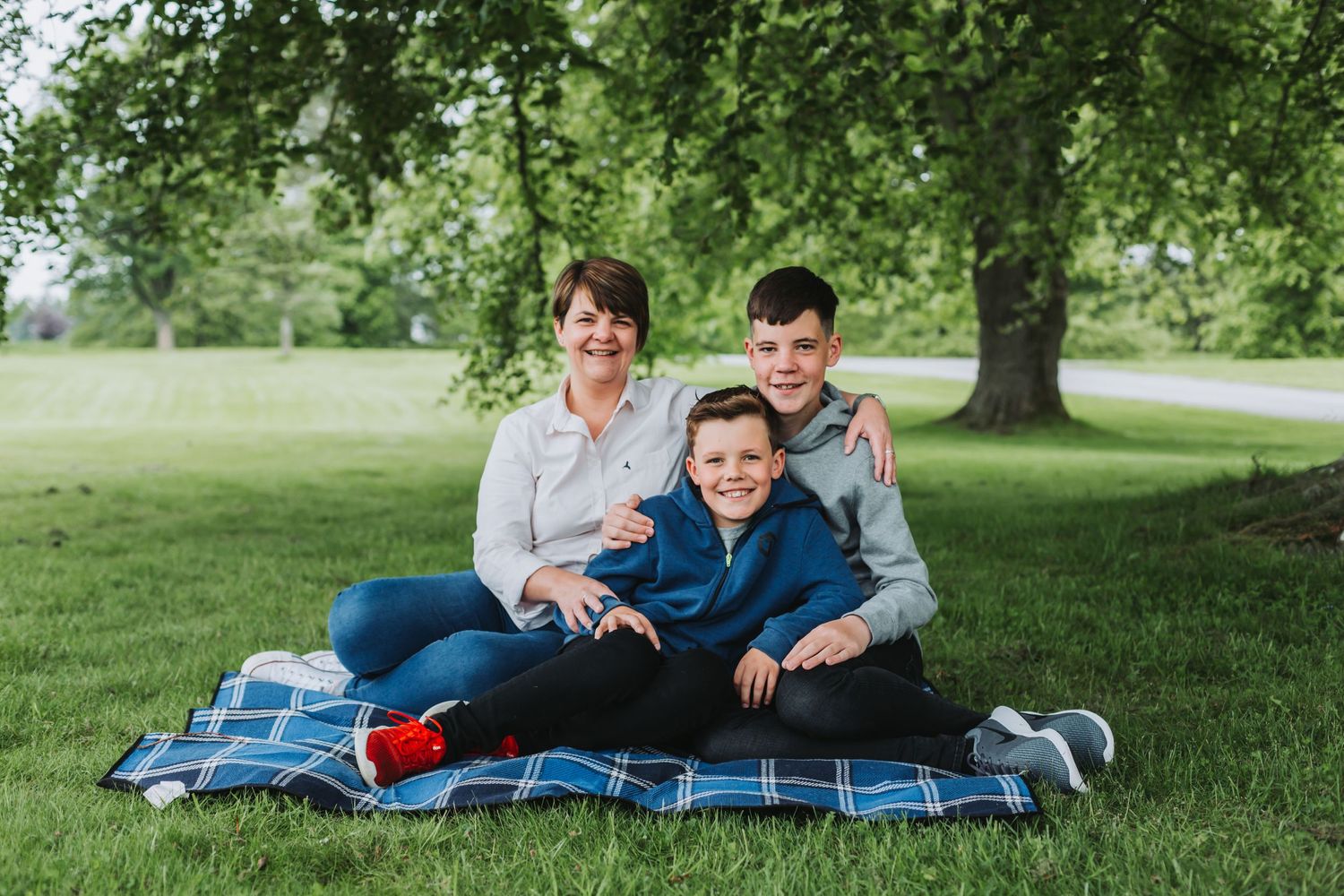 Woman and two boys smiling together on a plaid blanket under a large green tree in a park.