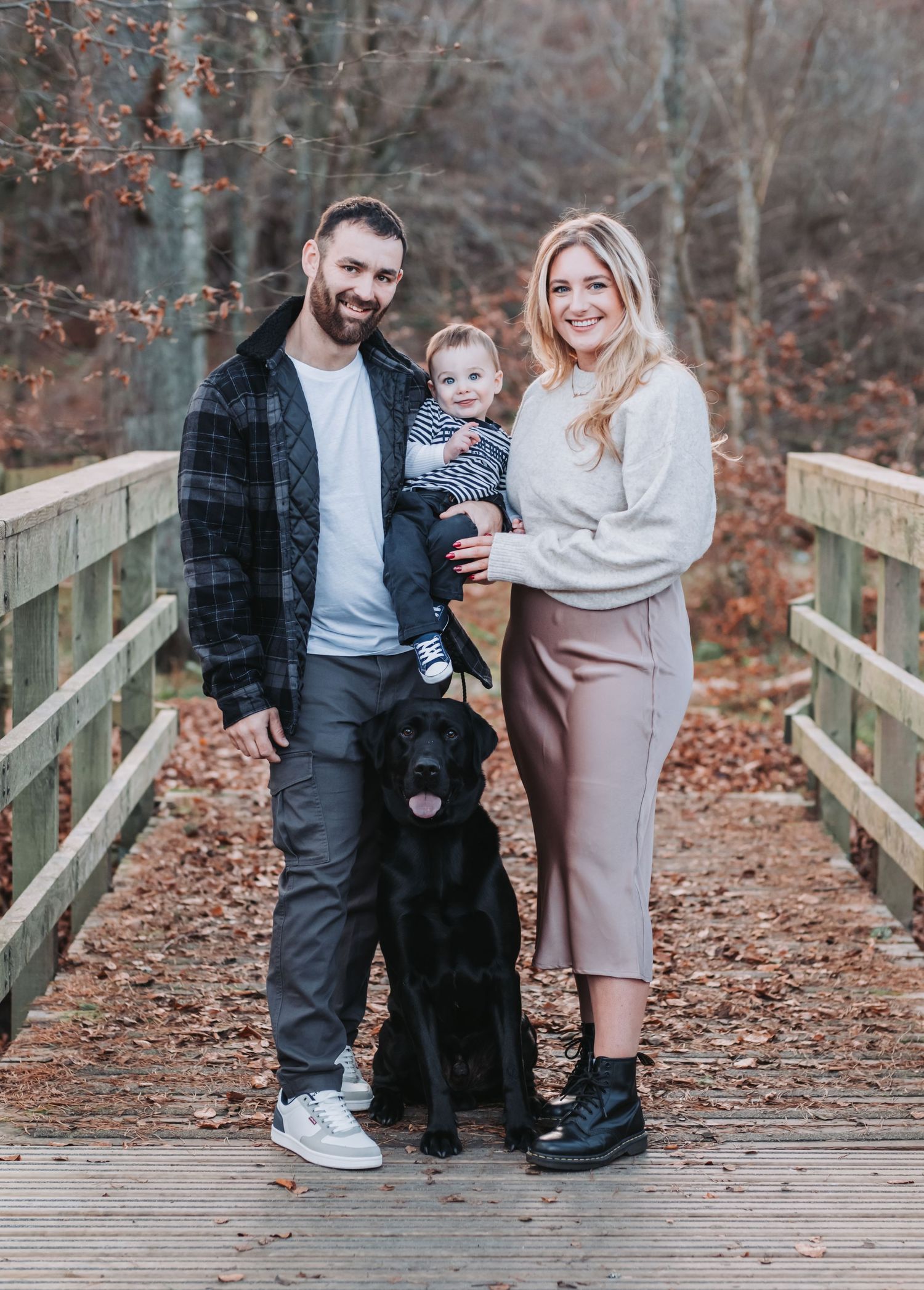 Smiling family with baby and black Labrador dog posing on a wooden bridge in autumn forest.