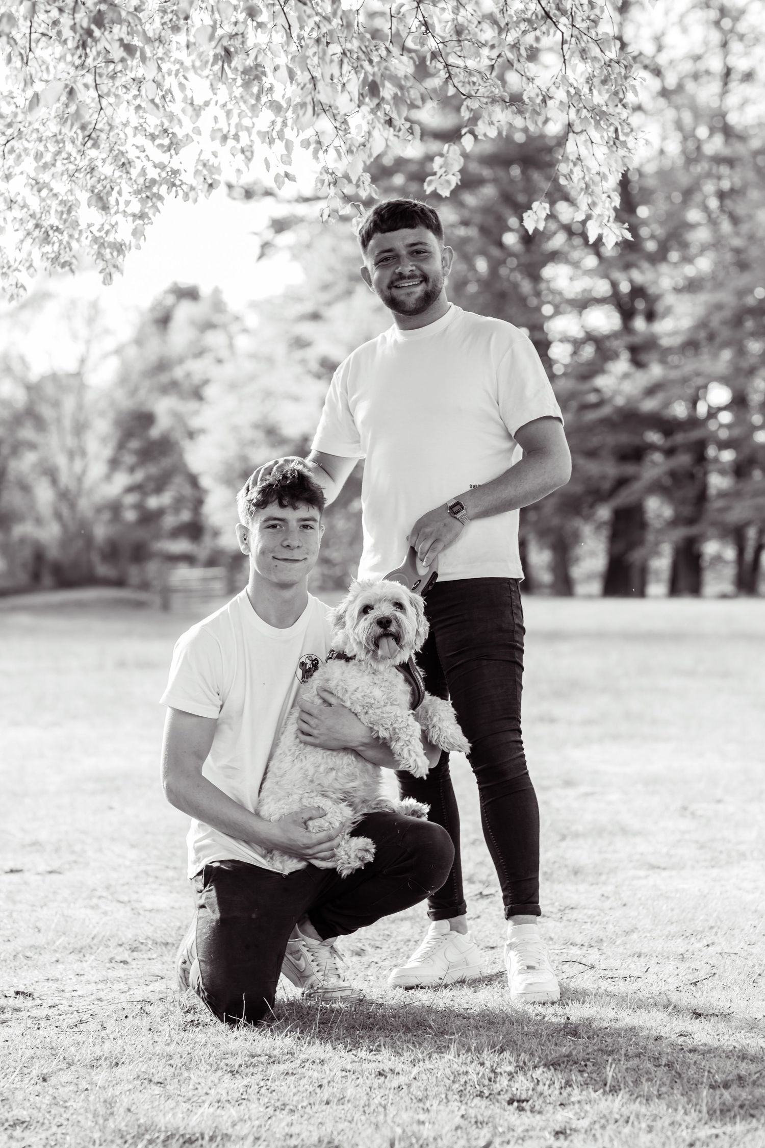 Black and white portrait of two young men posing outdoors in a park, one holding a fluffy dog.