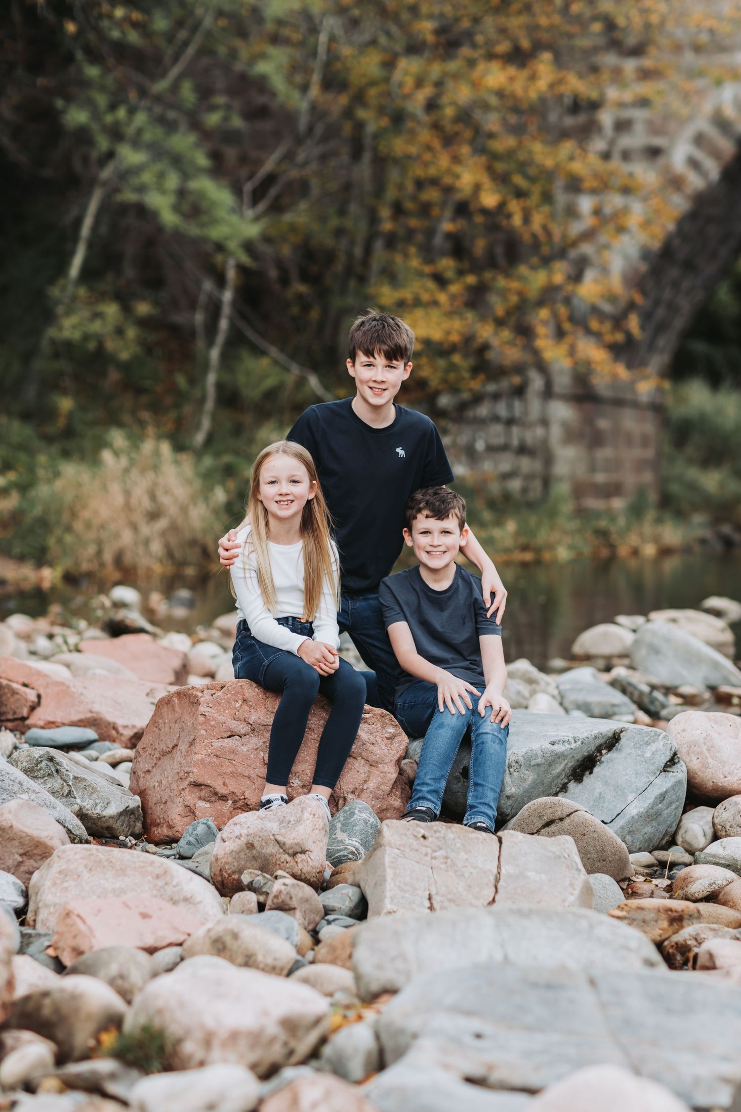 Three siblings smiling together while sitting on large rocks near an autumn riverside bridge.