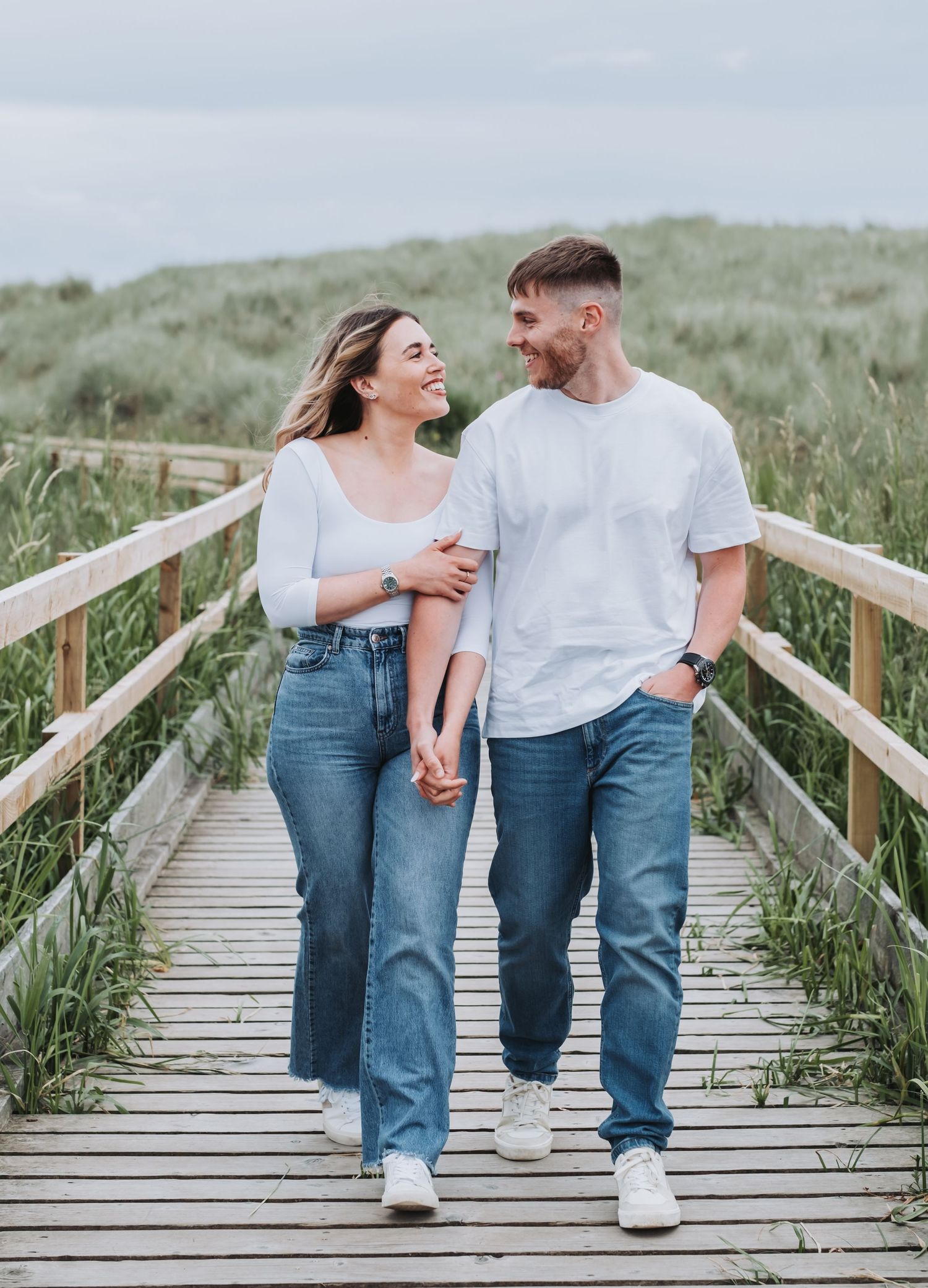 Young couple in matching white tops and jeans walking hand in hand on a wooden boardwalk.